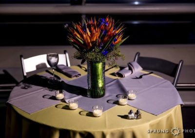 Sweetheart table for two with yellow and gray linens and votive candles. A bouquet of spiky orange and smaller dark blue flowers in a green vase sits in the middle of the table.