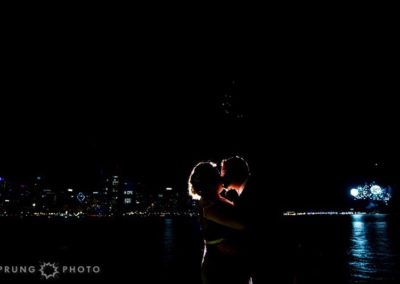 Two people kissing outdoors at night with fireworks and the lights of the Chicago skyline behind them.