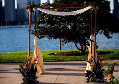A chuppah (canopy) made from four dark poles and a lace tablecloth. The front 2 poles have large arrangements of tropical flowers in front of them. In the background is a tree, some water, and buildings beyond the water.