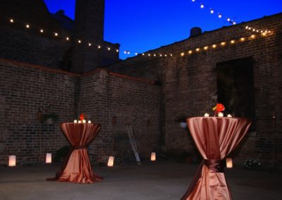 Two highboys draped in shiny brown linens set in a brick-walled courtyard. Luminaria line the edges of the space and there are strings of lights overhead.