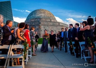 A bride, wearing a long, gray dress and carrying a bunch of spiky orange flowers and escorted by her mother, walks down the aisle at an outdoor wedding ceremony toward the camera. Wedding guests on either side, stand and turn toward them. In the background is a stone building with a dome.