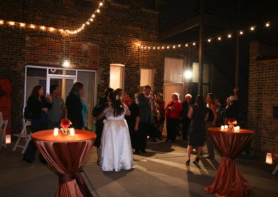 Wedding guests in a brick-walled courtyard. Two highboys draped in shiny brown fabric are in the foreground. Strings of lights are overhead.