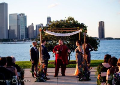 A marriage ceremony under a chuppah. The officiant stands in the middle, wearing a red tunic and pants. The groom and best man are on the left. The bride and maid of honor are on the right. Behind them are a tree, an expanse of water, and part of the Chicago skyline.