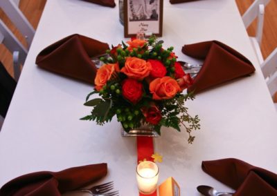 A long banquet table seen from one end. It has a white tablecloth, brown napkins, an orange ribbon down the middle, 2 centerpieces of orange roses, and candles.