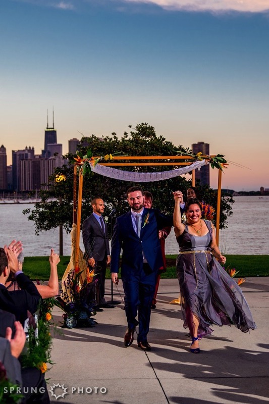 A man and a woman (wearing a blue suit and a gray dress, respectively) walk toward the camera and away from a chuppah decorated with lace and flowers. They raise their joined hands and people seated on either side are seen applauding. In the background are Lake Michigan and the Chicago skyline.