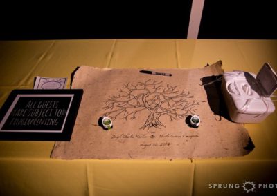 A large piece of handmade paper with an ink drawing of a bare tree on it. A sign to the left reads, "All guests are subject to fingerprinting." Small ink pads, a pen, and a box of wipes surround the paper. Two names and a date are written at the bottom of the paper.