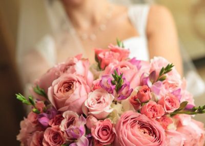 In the foreground, a round bouquet of pink roses of various sizes and shades, held by the woman in the background (out of focus) who is smiling and wearing a white dress and veil.