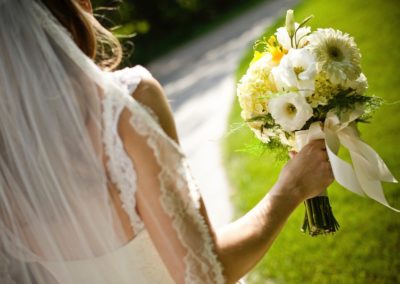 The torso of a woman wearing a white dress and veil, seen from behind. She is holding a bouquet of yellow and white flowers tied with a white ribbon.