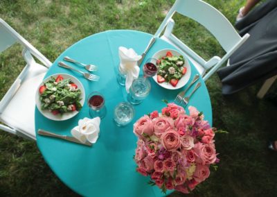 Overhead shot of a table for two. It has an aqua linen and white napkins in the water glasses. Two salads are served. It is decorated with a round bouquet of pink roses in various sizes and shades.