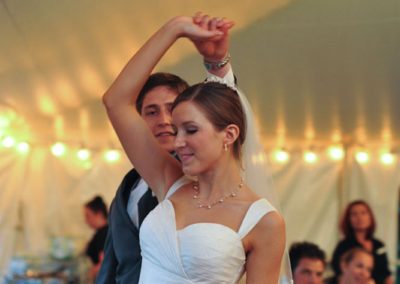 A man in a grey suit and a woman in a white dress and veil dance. He is twirling her.
