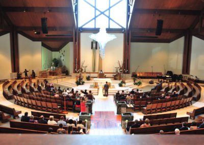 View from the balcony of a wedding ceremony in a modern-looking church. The seating is in a semi-circle around the altar, and there is a large, clear window behind the altar. The seating, floor, and ceiling are dark wood tones, while the walls are white.