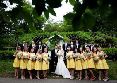 Portrait of a wedding party. The groom, in a dark suit, and the bride, in a long white dress and veil, stand in the center. The 11 bridesmaids (in front) and 11 groomsmen (behind) stand on both sides of them. The men wear dark suits; the women wear knee-length yellow dresses and hold bouquets of white flowers.