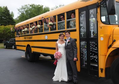 A woman in a white dress and veil carrying a bouquet and a man in a grey suit stand in front of a yellow school bus which is decorated to say "Just Married." Their wedding party looks out of the windows of the bus.