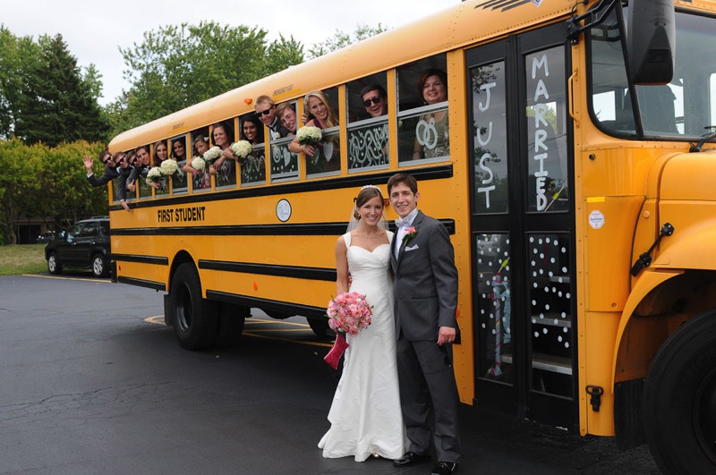 A woman in a white dress and veil carrying a bouquet and a man in a grey suit stand in front of a yellow school bus which is decorated to say "Just Married." Their wedding party looks out of the windows of the bus.