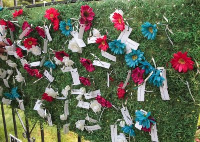 A piece of green outdoor carpet hanging on a fence. Attached to the carpet are red and blue silk flowers, each one attached to a place card.