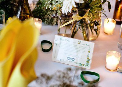In focus is a mason jar containing a few white flowers and some greenery. Out of focus are two yellow napkins in water glasses, votive candles, and other table decor.