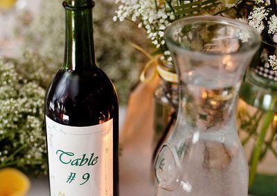 A wine bottle whose label says, "Table #9" next to a carafe of water and a a mason jar containing ferns, daisies, baby's-breath and other flowers.