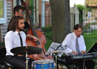 A jazz trio playing outdoors: drummer, bassist, and keyboard player.