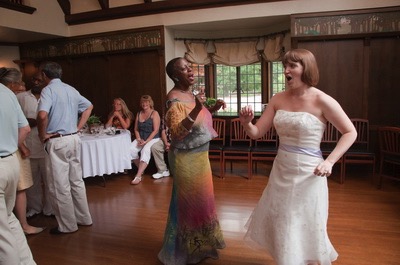 Two women dancing in a wood-paneled room. One wears a white dress; the other wears a multi-colored dress.