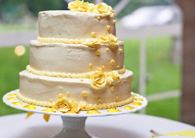 A home-made, 3-tier wedding cake on a cake stand covered in white frosting and decorated with yellow sugar-flowers. It is framed in the arched window of a tent.