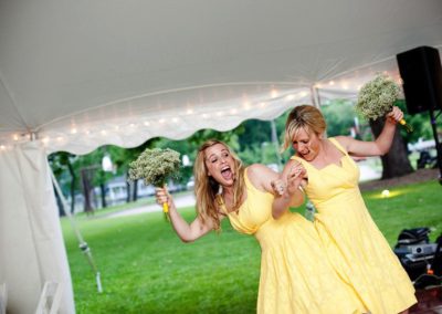 Two women wearing knee-length yellow dresses and carrying bouquets of white flowers dance at the entrance to a tent.