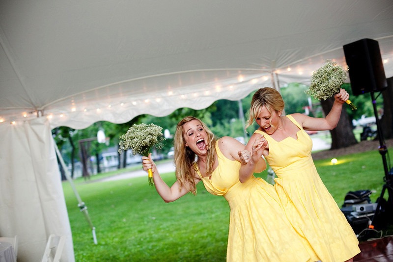 Two women wearing knee-length yellow dresses and carrying bouquets of white flowers dance at the entrance to a tent.