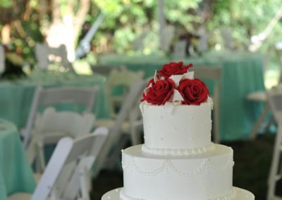 A white wedding cake topped with red roses on a table in a tent.