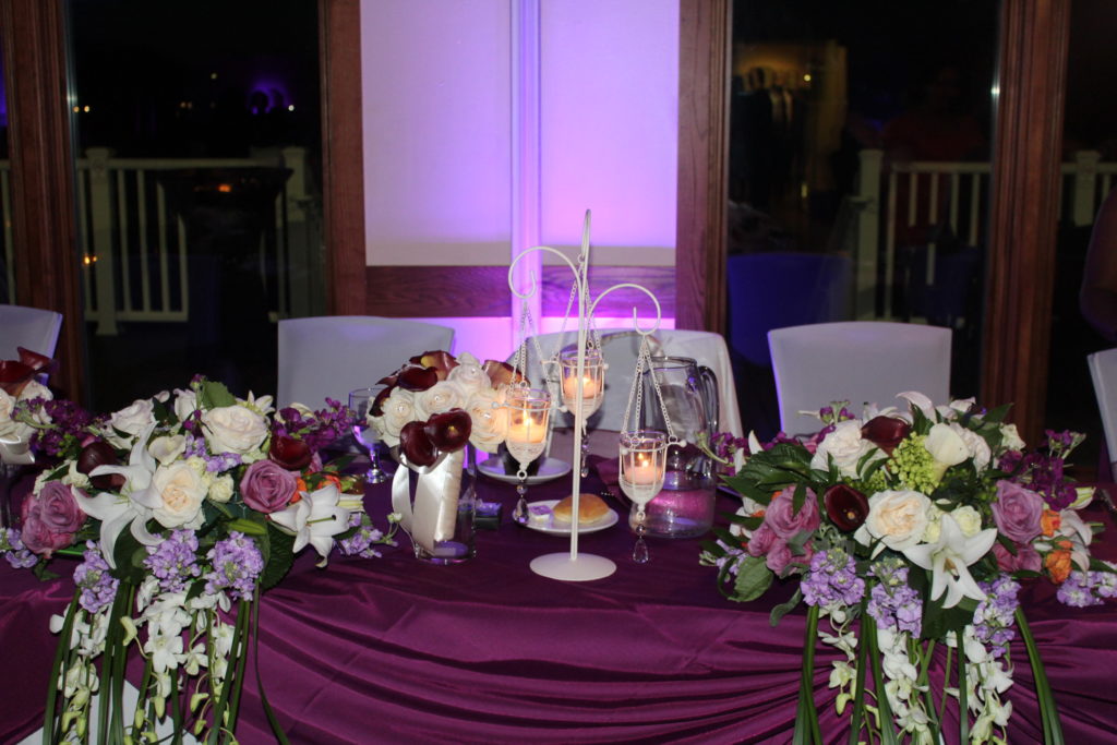 A table with a draped, deep purple cloth, a small chandelier, and two bunches of pink and purple flowers cascading over the front of the table.
