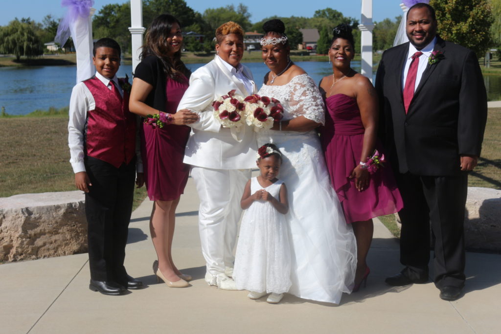 A beautifully dressed Black family in white, black, and burgundy, standing in a row and smiling, in front of a small lake.