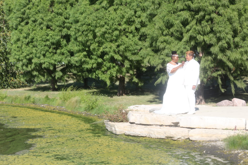 Two Black women in white, one in a wedding dress, the other in a suit, standing in front of green trees and next to a small lake.