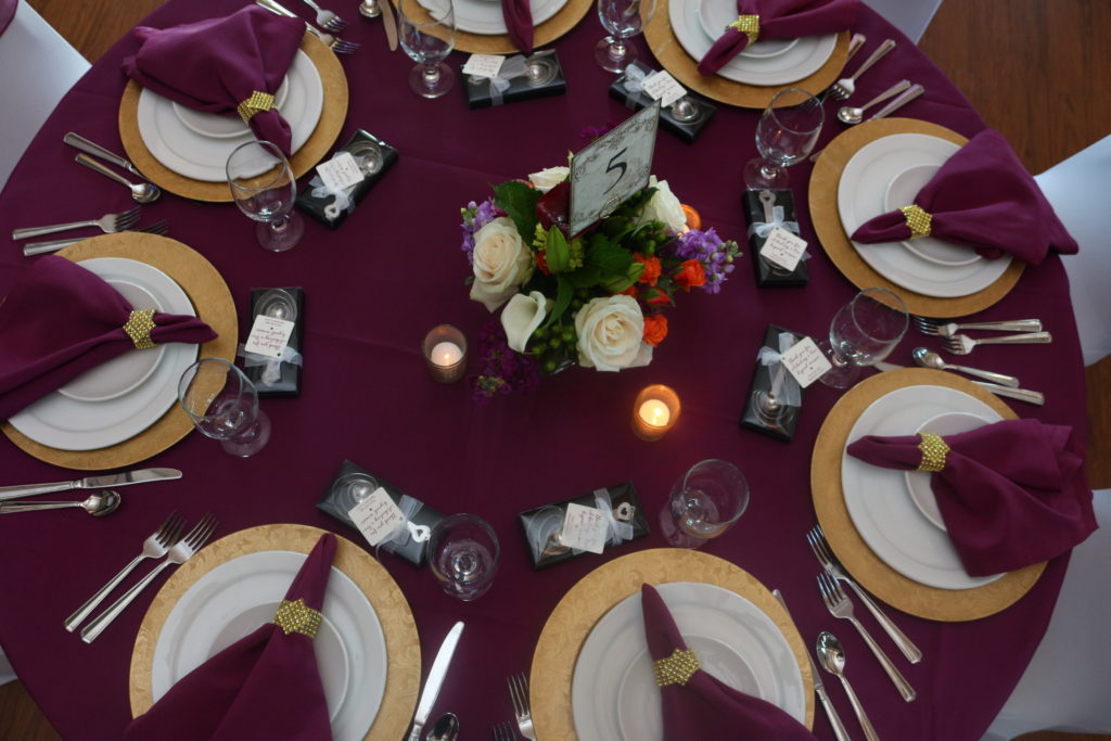 A round banquet table seen from overhead, with purple cloth and napkins, gold chargers, white plates, gold napkin rings, candles and pink and purple flowers in the center