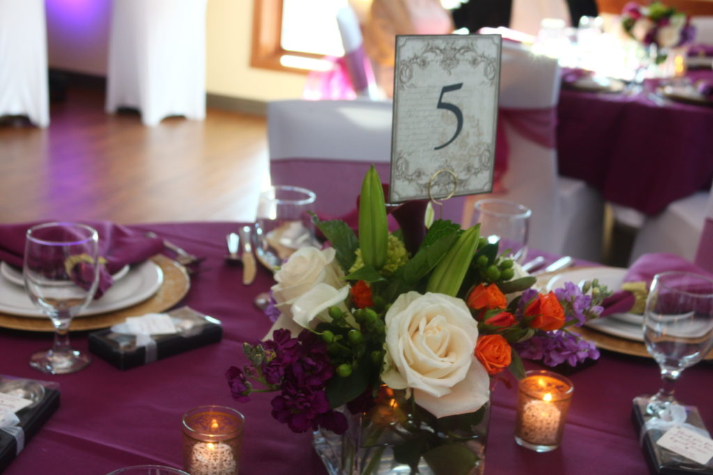 A floral centerpiece of pale pink, red, and purple flowers and a table number on a table with a purple cloth.
