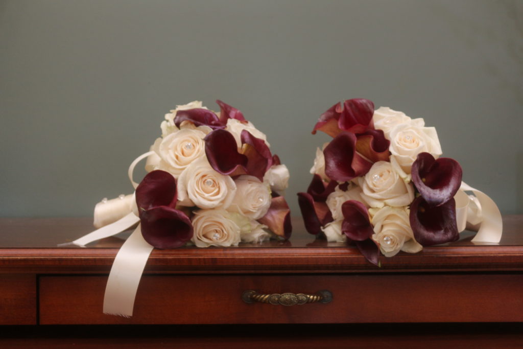 Two bridal bouquets on a wooden table.  Bouquets are of very pale pink roses and burgundy lilies, wrapped with pale pink ribbon.