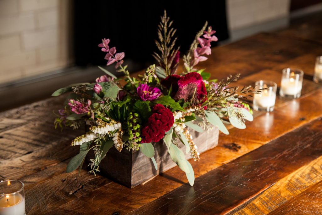 Red, pink, and white flowers in a rectangular wooden planter on a shiny wood farm table, flanked by votive candles.