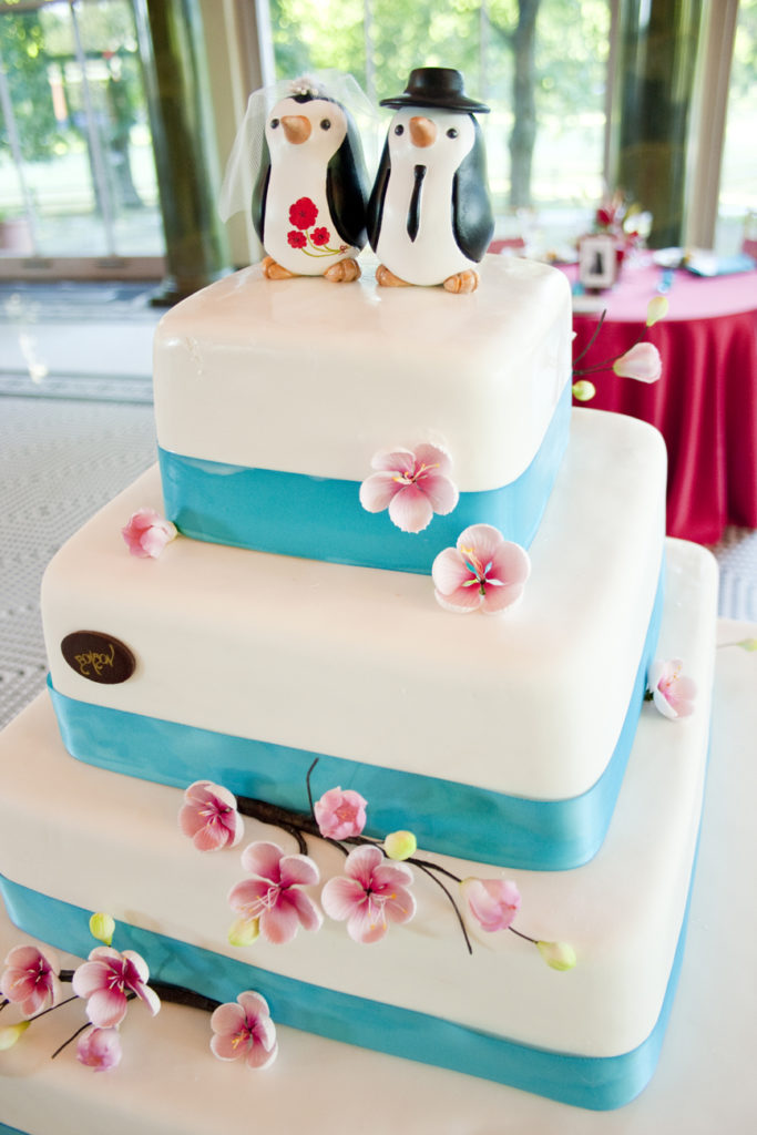 A square white wedding cake with blue ribbons around each layer, decorated with pink cherry blossoms and a pair of bride-and-groom penguins on top.