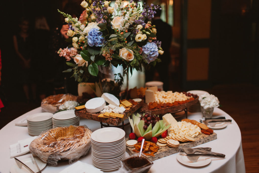 A spread of cheese and crackers on a round table dominated by a tall floral arrangement in peach, blue, and purple.