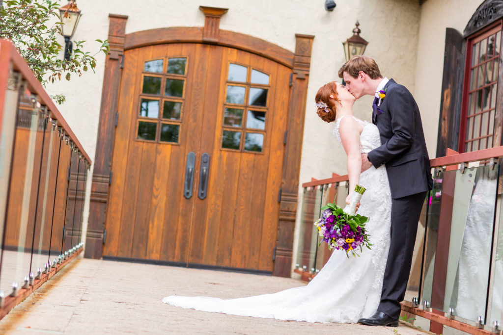 A woman in a white dress and carrying a bouquet and a man in a blue suit kiss each other in front of a pair of wooden doors.