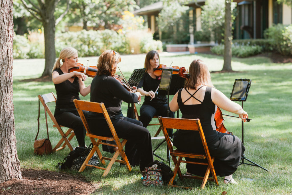A string quartet, sitting in wooden chairs outdoors, playing.