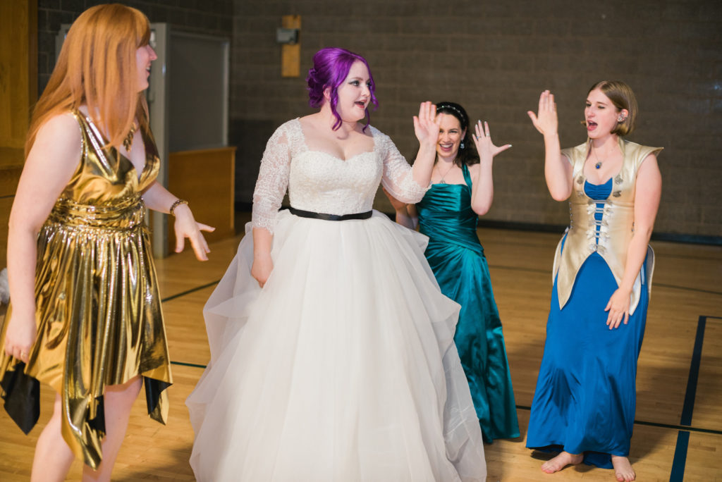A woman with purple hair in a white wedding dress with a black sash dances with three people in costume dresses.