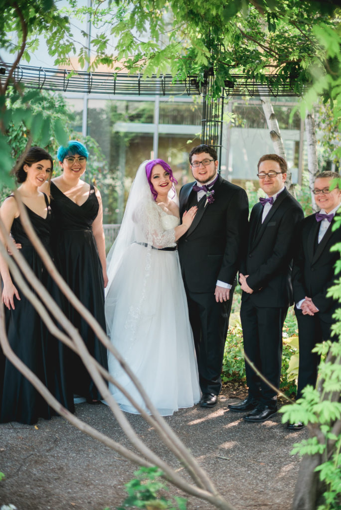 Photo of a wedding party outdoors. The bride has purple hair and wears white. Everyone else wears black, either long dresses or suits. 