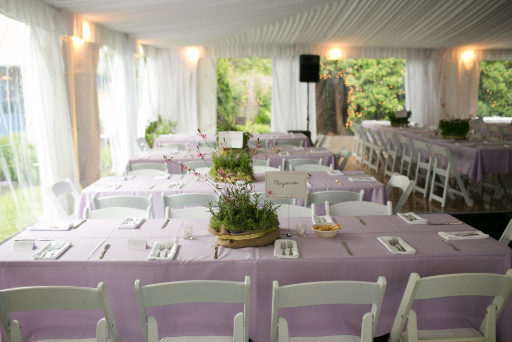 The interior of an elegant tent containing long tables with lavender cloths, white chairs, and centerpieces of herb plants in burlap.