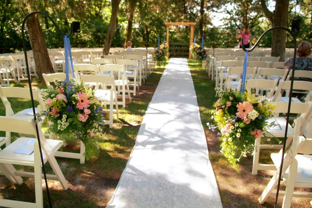 A view down the aisle for a wedding ceremony. A white runner goes the length of the aisle. White chairs in rows are on either side and a square wooden arch is at the far end. Flowers hanging from hooks frame the foreground. Some bare dirt is seen on either side of the runner.