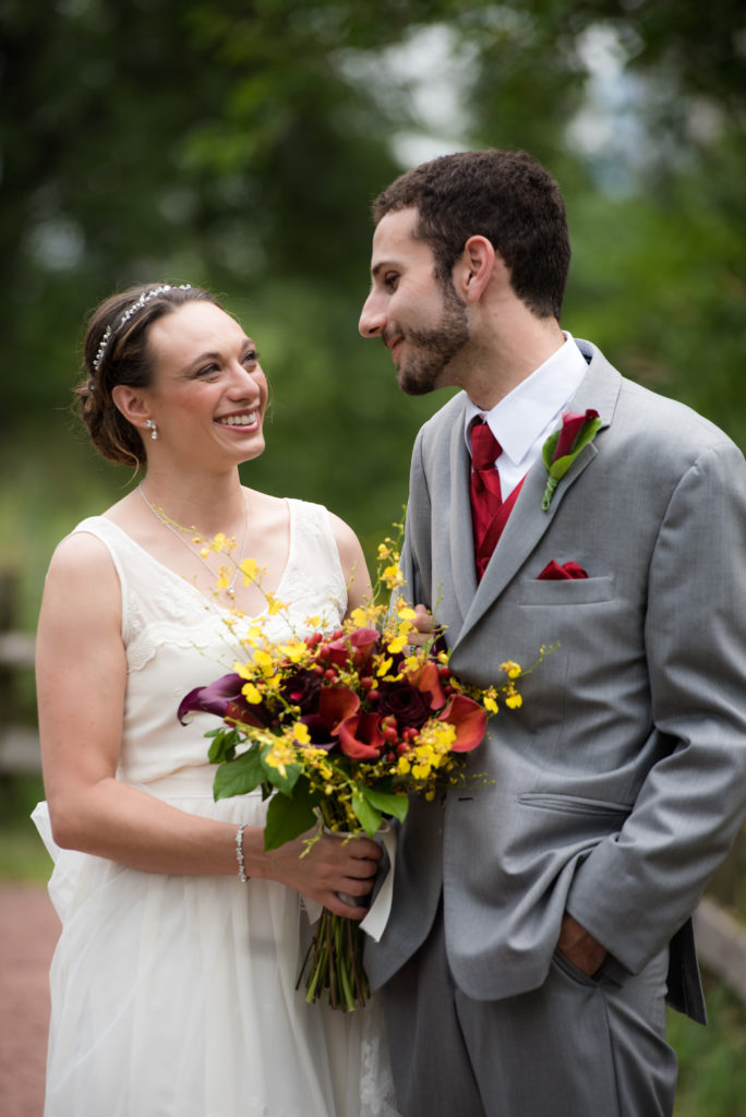 A bride and groom looking at each other. The bride wears white and carries a bouquet of orange, red, purple, and yellow flowers. The groom wears a gray suit with a red tie and hankerchief, and a red boutonniere. The background is outdoorsy.