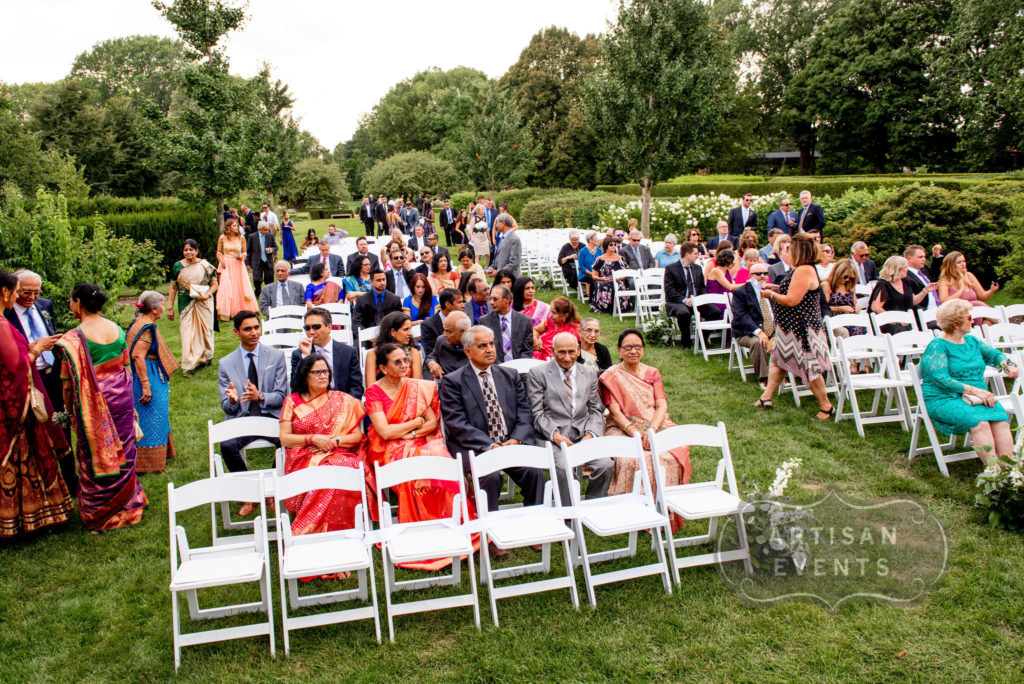 The guests at an outdoor wedding ceremon are seated in rows in a green garden. Many are in Indian dress.