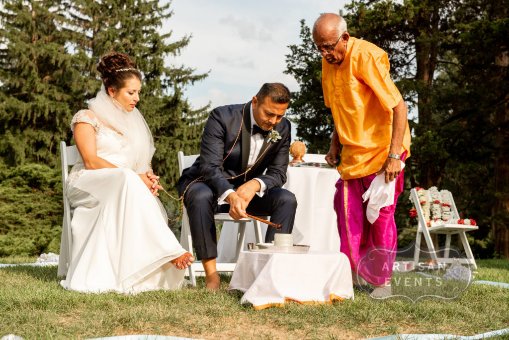 Bride and groom are seated for part of a Hindu marriage ceremony. A Hindu priest stands to their left.