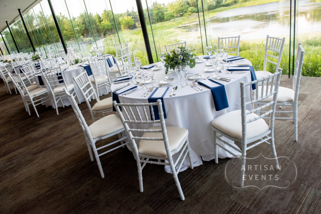 A row of round banquet tables with white linen, navy blue napkins, and floral centerpieces. Outside the windows in the background are a pond and trees.