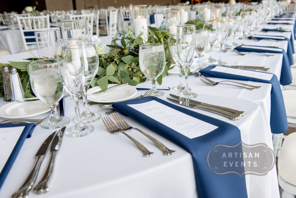 A long table with a white linen and navy blue napkins, deecorated with a long garland of greenery and pillar candles.