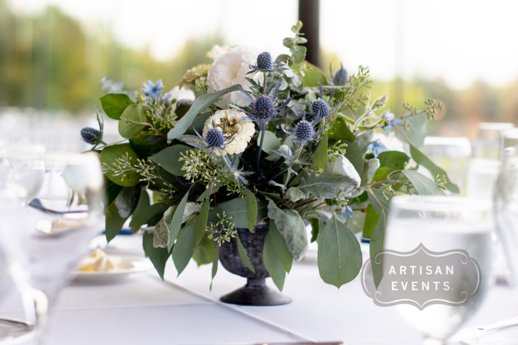 A rounded floral centerpiece in a pedestal bowl, comprised of greenery, white and pale pink blossoms, and blue thistle flowers.