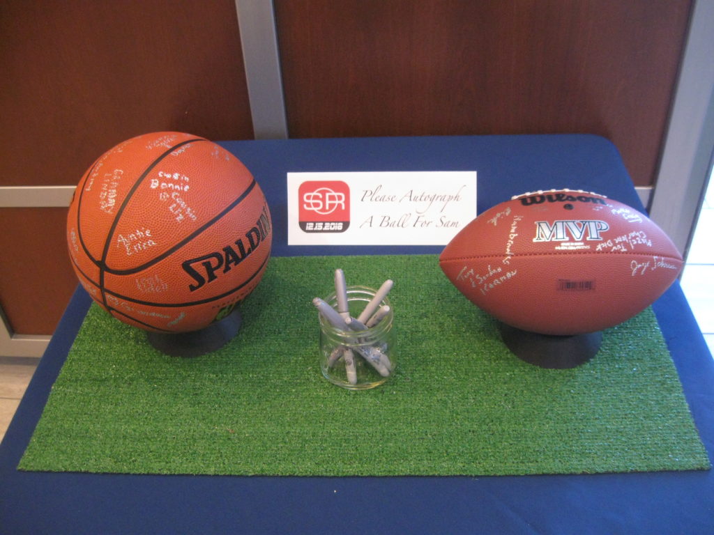 A basketball and a football on a table covered in astroturf. A jar of pens is between them, with a sign that says, "Please autograph a ball for Sam."