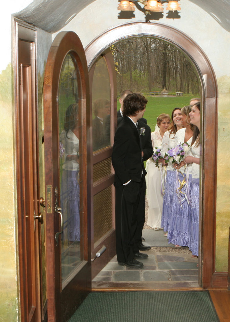 An arched doorway seen from inside. Outside a wedding party waits to come in. Greenery is in the distance.